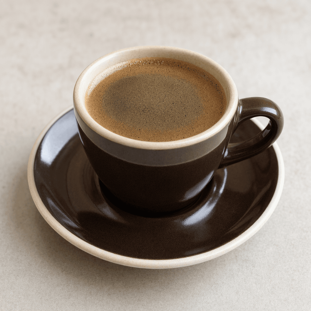 A small cup of dark brown coffee with a smooth surface and a thin layer of tan foam, served on a saucer.