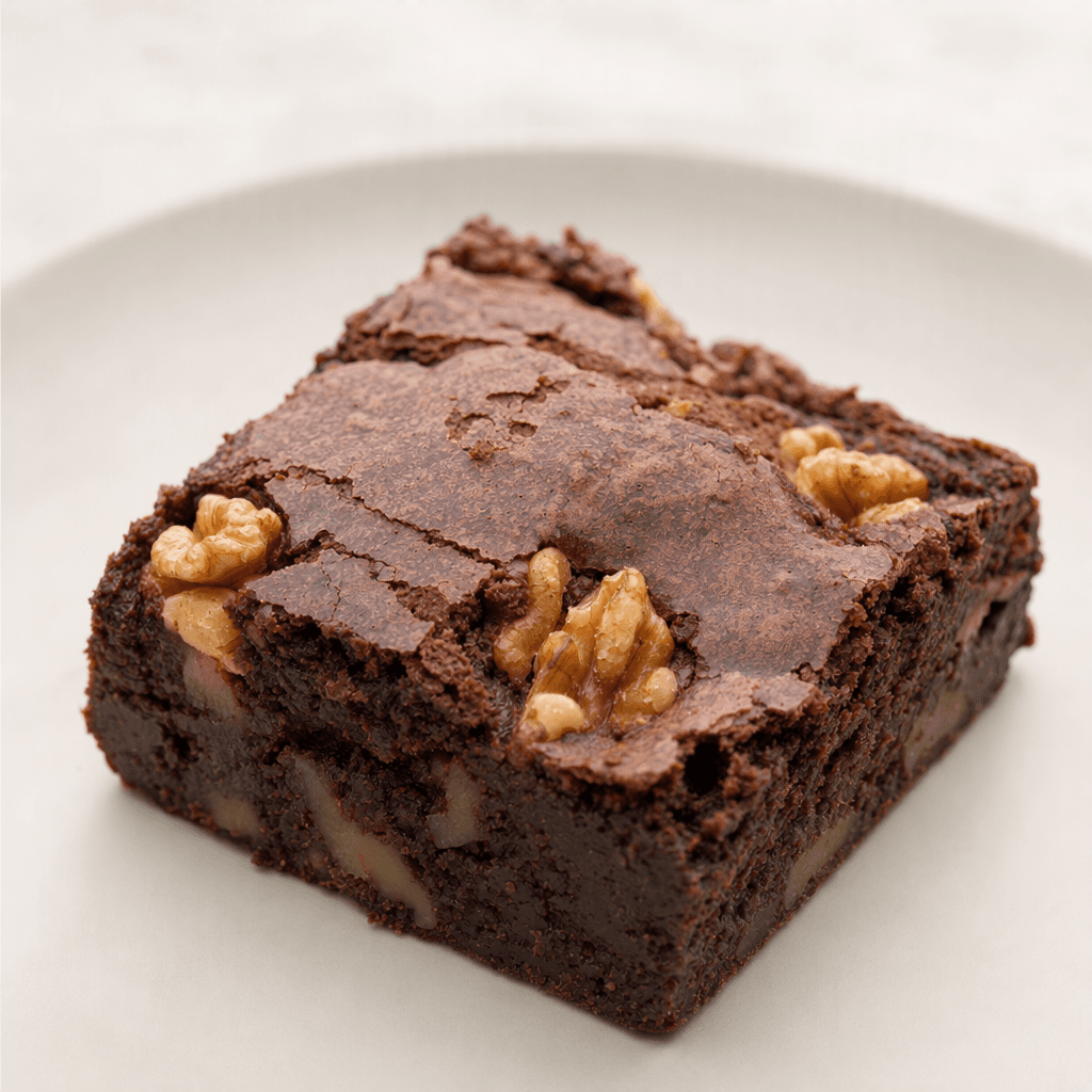 Üstü hafif çatlamış, içindeki iri ceviz parçaları görünen koyu kahverengi, yoğun dokulu kare bir brownie sade bir tabakta servis ediliyor.
