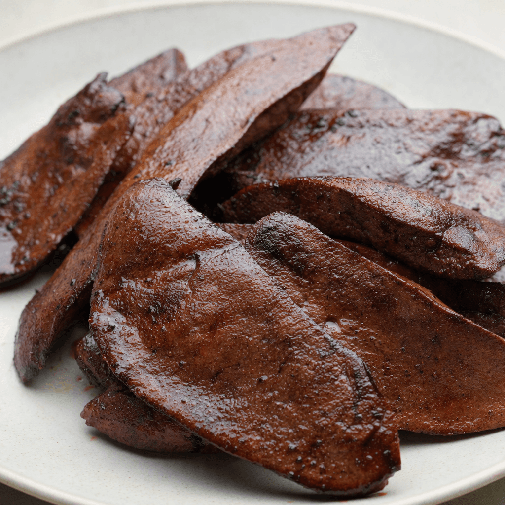 Several irregular slices of grilled spleen with a deep reddish-brown surface, darker seared patches, and a dense, slightly glossy texture are arranged on a plate.