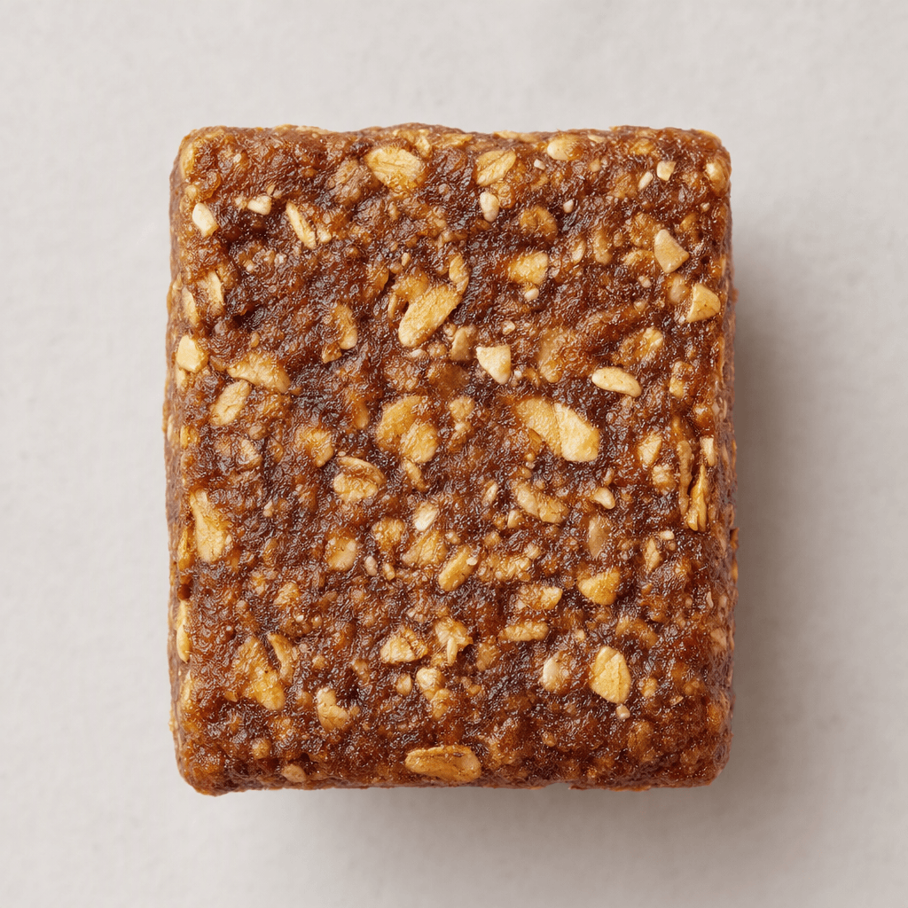 A compact, rectangular brown bar with a coarse, slightly uneven surface and visible pale oat flakes throughout.