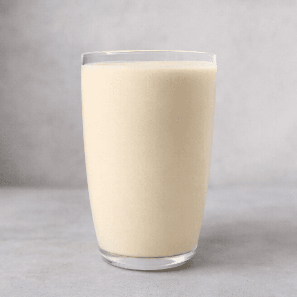 A smooth, opaque, pale cream-colored nutritional drink sits in a clear glass, with an even surface and no visible pieces.