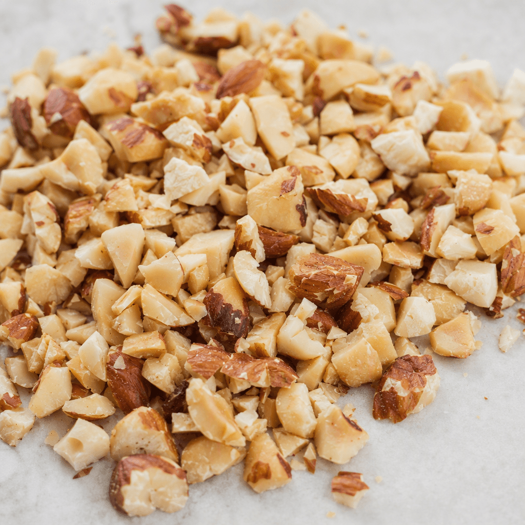 Small, irregularly chopped hazelnut pieces with warm tan to light brown interiors, patches of darker papery skin, rough crumbly surfaces, and varied angular shapes, scattered in a loose pile.