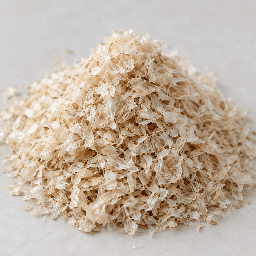 Loose pale beige psyllium husks form a small mound, made of thin, curled, translucent papery flakes with an airy, fibrous texture.