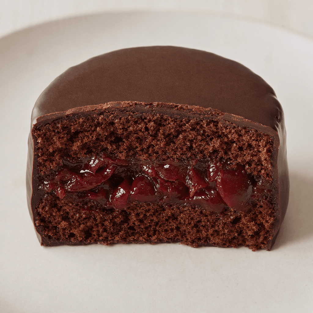 A small, round cocoa sponge cake with a dark chocolate coating and a glossy red sour-cherry filling visible in the cut center, shown on a plain plate.