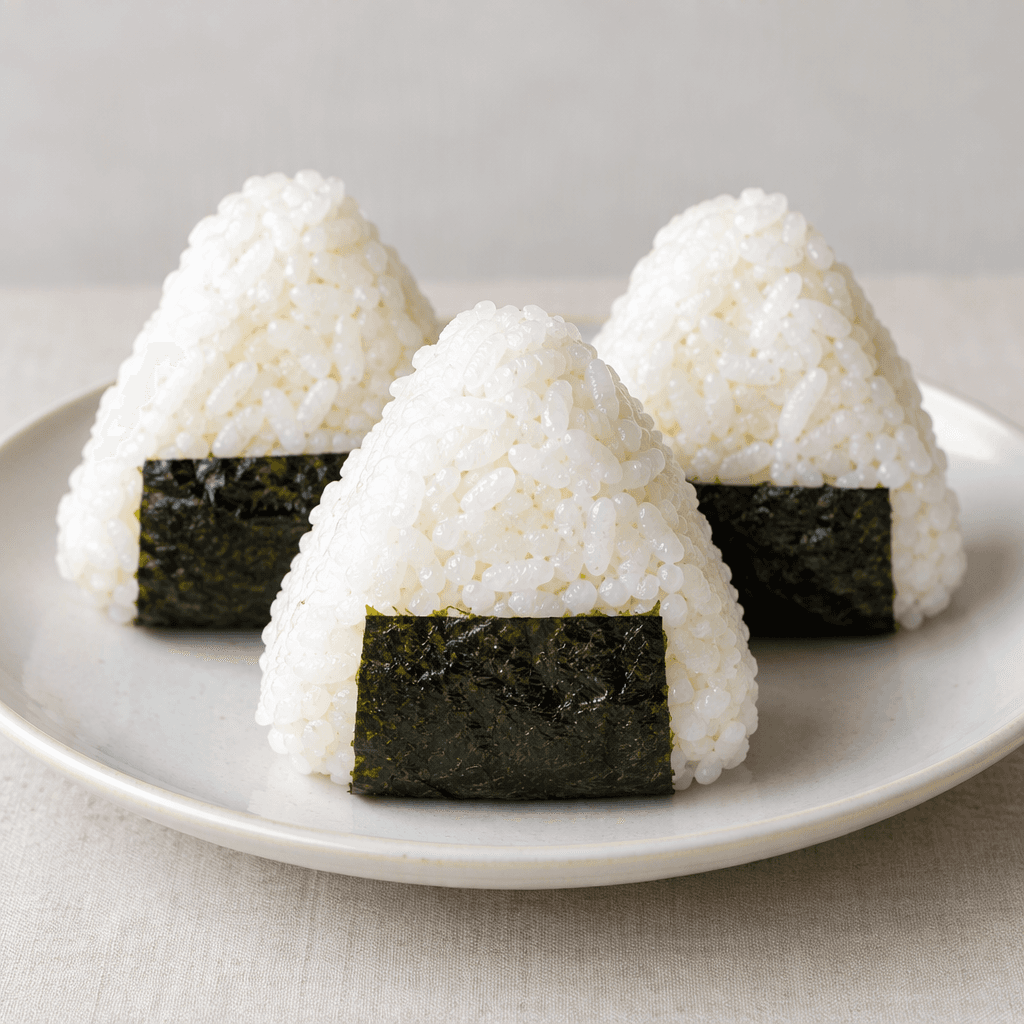 Three compact triangular mounds of white, slightly glossy rice are arranged on a simple plate, each wrapped with a dark green-black strip of nori along the base and showing softly uneven, grain-textured surfaces.