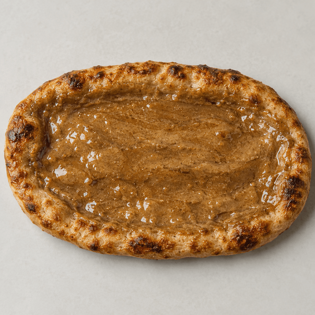 An elongated oval flatbread with a thin, blistered golden-brown crust, a dark beige glossy tahini-and-sugar layer spread across the surface, and slightly raised, deeply browned edges.