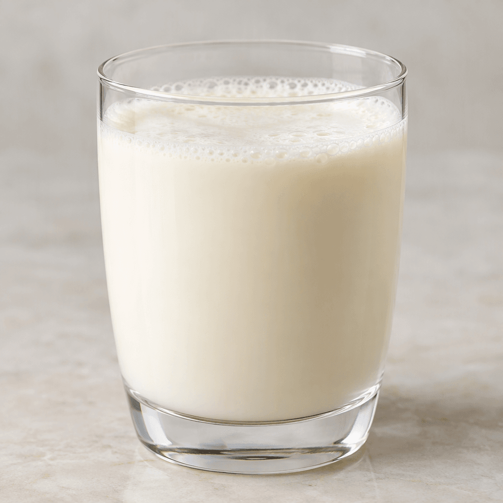 An opaque, pale ivory liquid with a smooth, lightly creamy appearance, poured into a clear glass with a thin layer of bubbles on top.