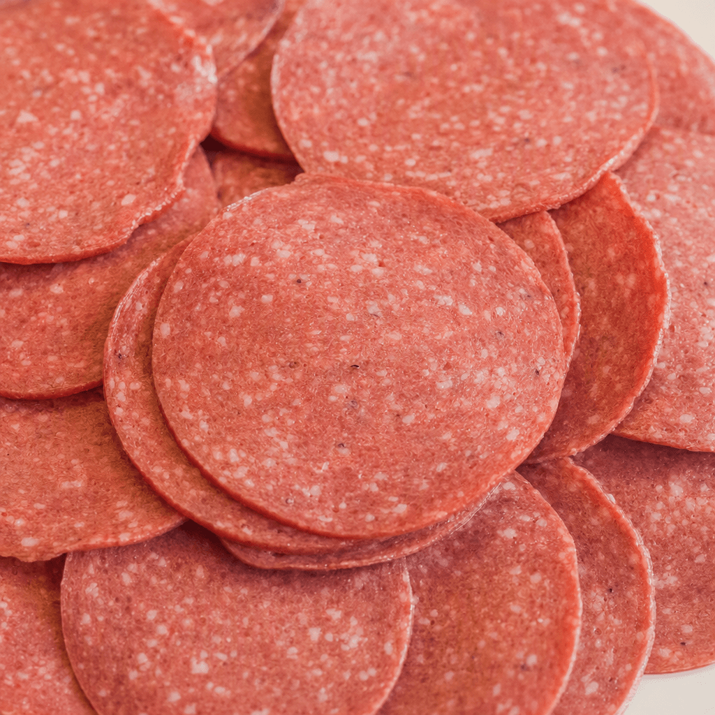 Thin, round slices of reddish-pink processed meat overlap in a loose stack, with small pale flecks scattered through their smooth, moist-looking surfaces.