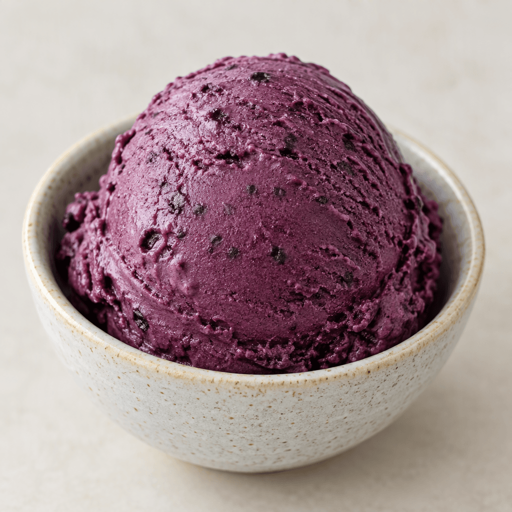 A rounded scoop of black mulberry ice cream, deep purple with a smooth, creamy surface and occasional darker berry flecks, served in a small bowl.