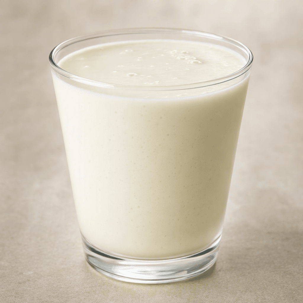 A pale white, smooth, slightly thick dairy drink sits in a clear glass, with a few tiny bubbles visible on its surface.