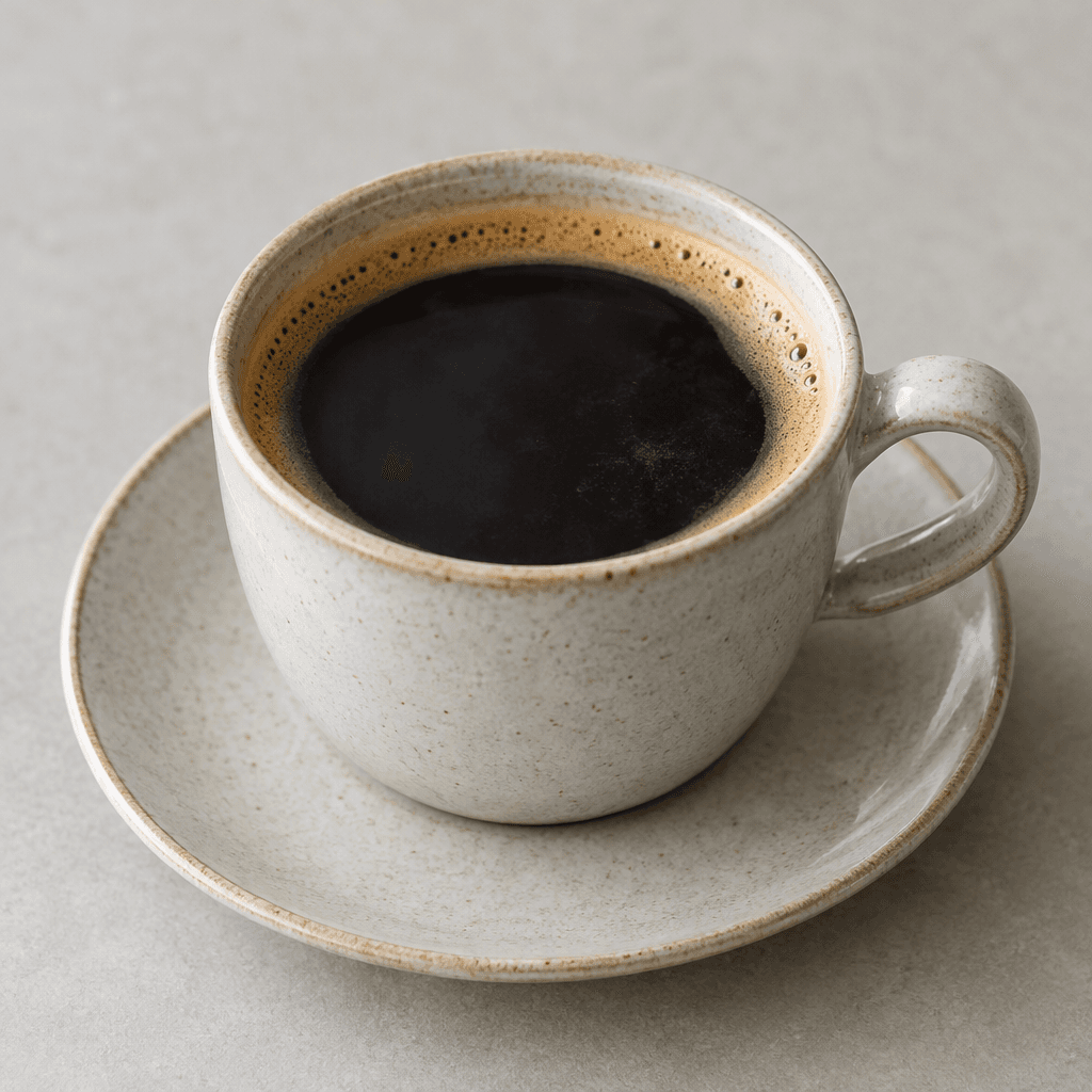 A ceramic cup holds a clear, deep brown-black coffee with a thin tan crema floating on the surface, set on a matching saucer.