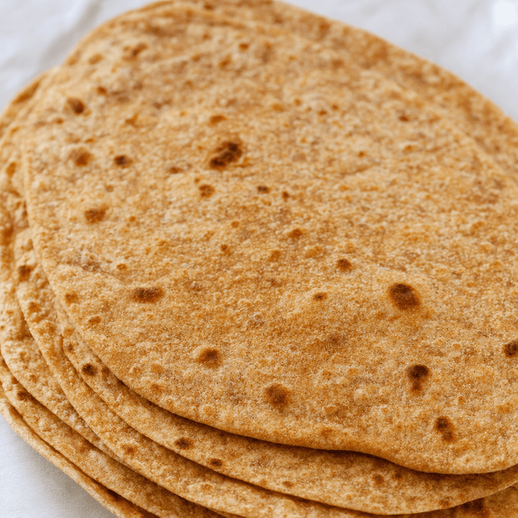 Thin, flexible oval sheets of whole-wheat flatbread, tawny brown with fine bran flecks and scattered darker toasted blisters, stacked slightly overlapping.