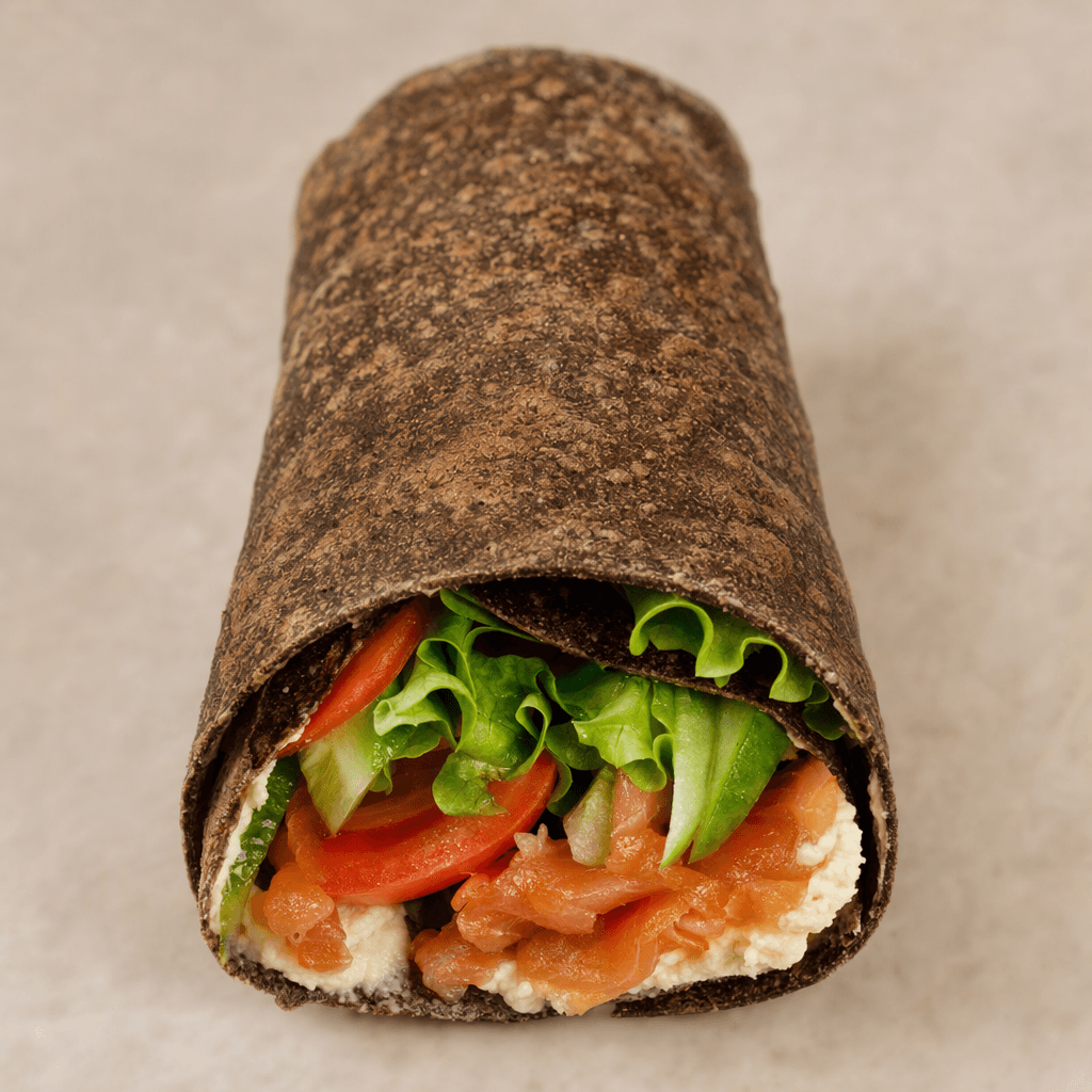 A thin, dark-brown rye flatbread with a lightly speckled, slightly uneven surface is rolled around a filling, with one end left open.