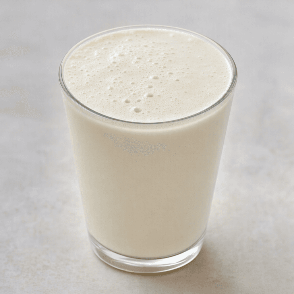 A smooth, opaque, pale cream-colored protein drink with a lightly foamy surface, served in a clear glass.