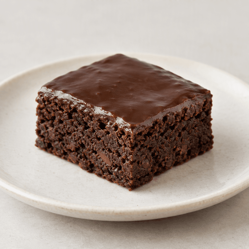 A small, dark-brown rectangular brownie with a dense, slightly uneven crumb and a smooth, glossy chocolate-coated top, served on a plate.
