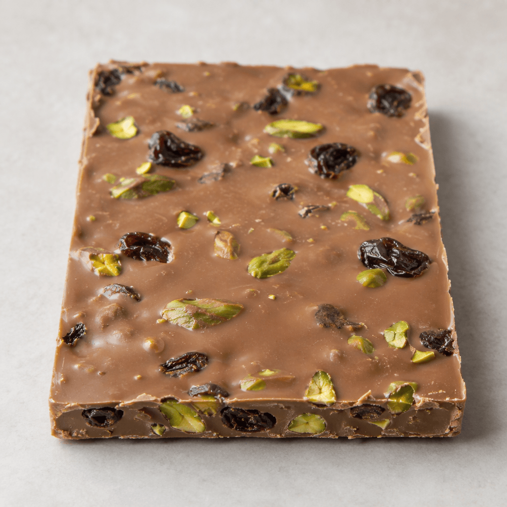 A flat, rectangular slab of pale milk chocolate with a smooth, slightly glossy surface, dotted with dark raisins and bright green pistachio pieces, some visible along the broken edges.