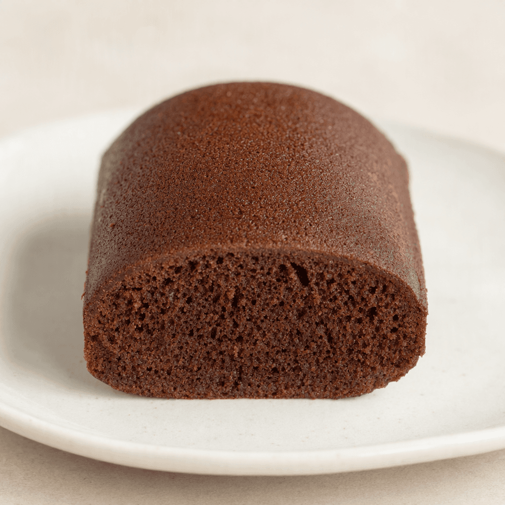 A small, rounded rectangular cocoa cake sits on a plate, with a dark brown, softly domed top and a porous, lighter-brown crumb visible along one cut edge.