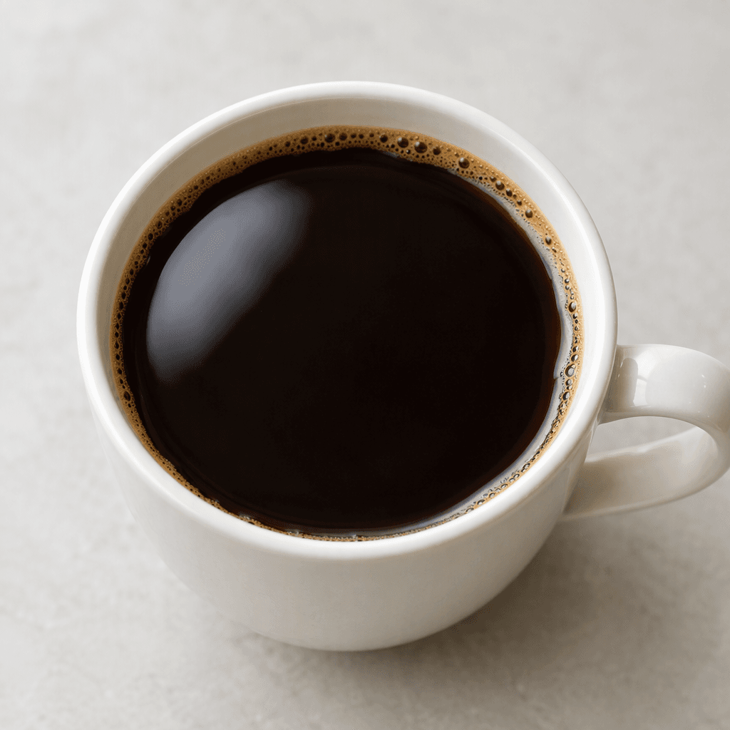 A cup filled with deep brown, nearly black brewed coffee, its smooth surface showing a soft reflective sheen and a thin ring of lighter brown foam along the edge.