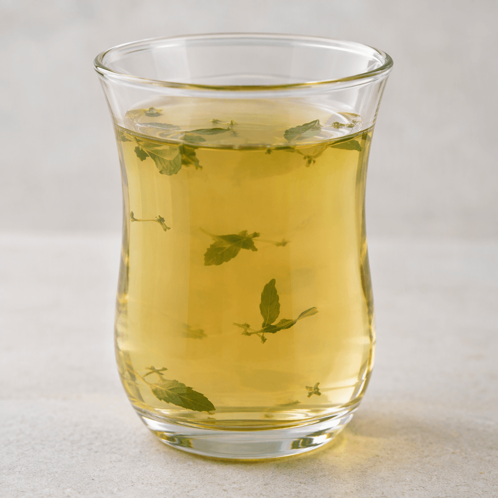 A translucent herbal infusion, pale amber-green in colour, sits in a clear glass with a few small green leaves visible in the liquid.