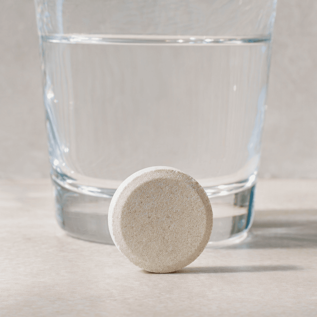 A pale, round, flat-sided effervescent tablet with a matte, finely porous surface sits beside a clear glass of water.