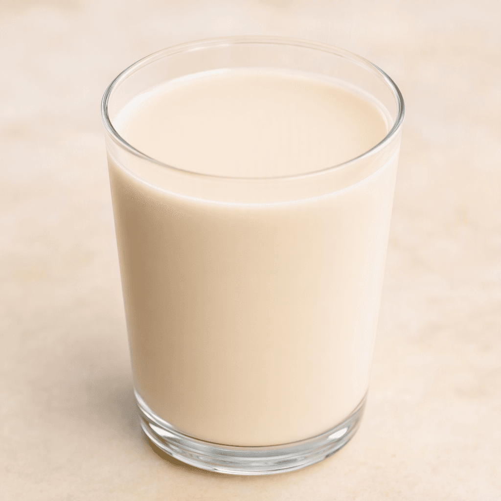 A smooth, opaque, pale beige milk sits in a clear glass, with an even surface and no visible foam.