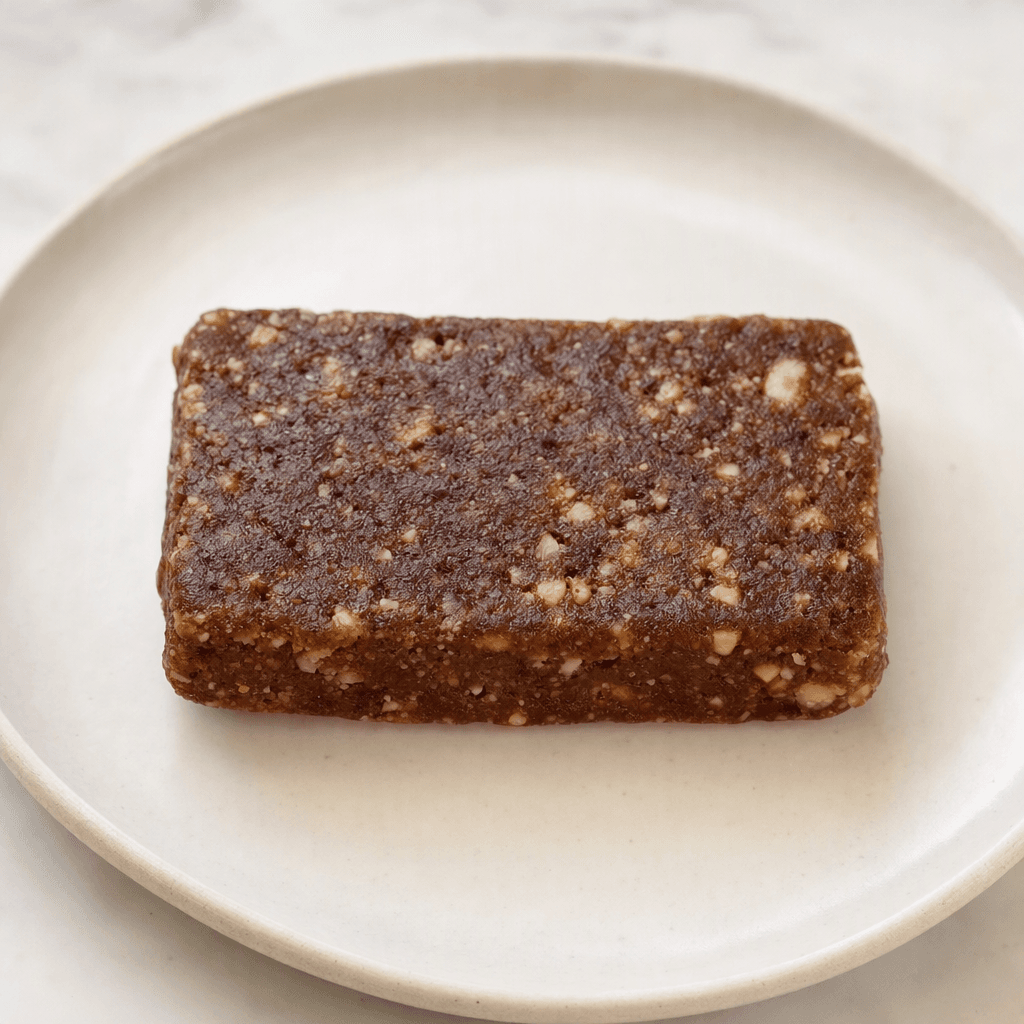 A compact, rectangular brown bar with a dense, slightly uneven surface and small pale nut or grain pieces visible throughout, served on a plain plate.