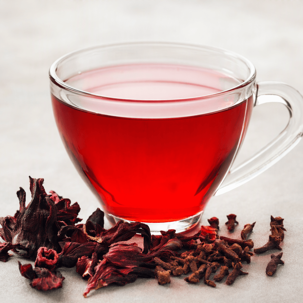A clear glass cup holds translucent deep ruby-red tea, with dark cloves and a few burgundy hibiscus petals visible beside it.