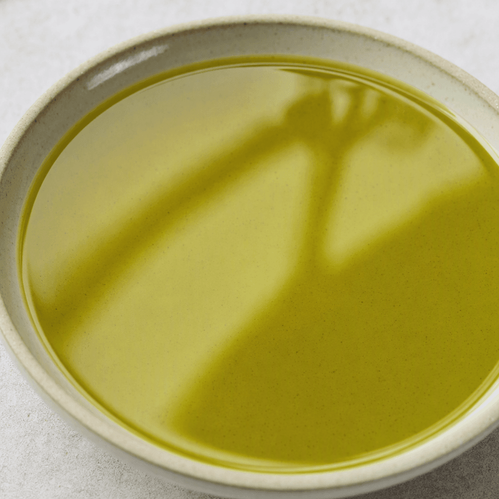 A clear, glossy, fluid oil with a golden-green hue, pooled in a shallow dish.