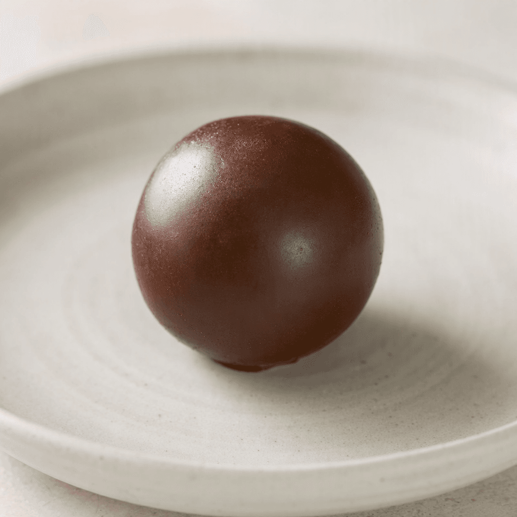 A small, round chocolate sweet with a smooth, dark-brown surface sits on a plate.