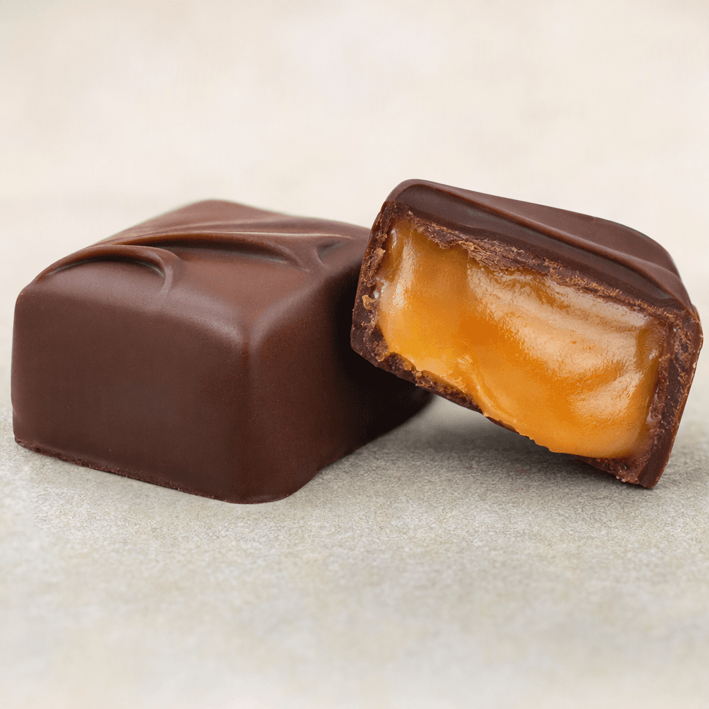 A dark brown chocolate piece with a smooth, slightly glossy shell is shown whole beside a cut piece revealing thick orange-amber caramel filling.