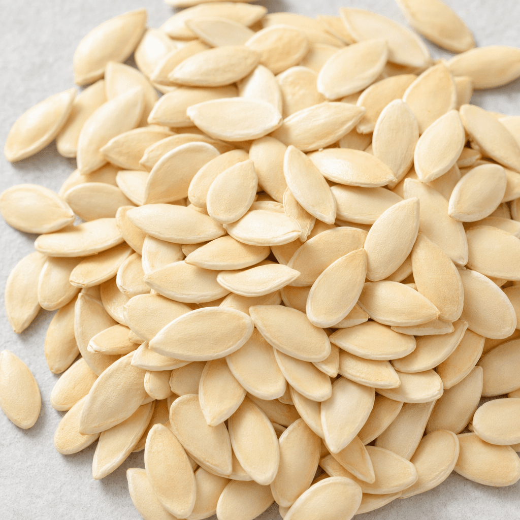 Small, flat, elongated oval melon seeds with pointed ends and smooth, pale cream-to-beige surfaces lie scattered in a loose pile.