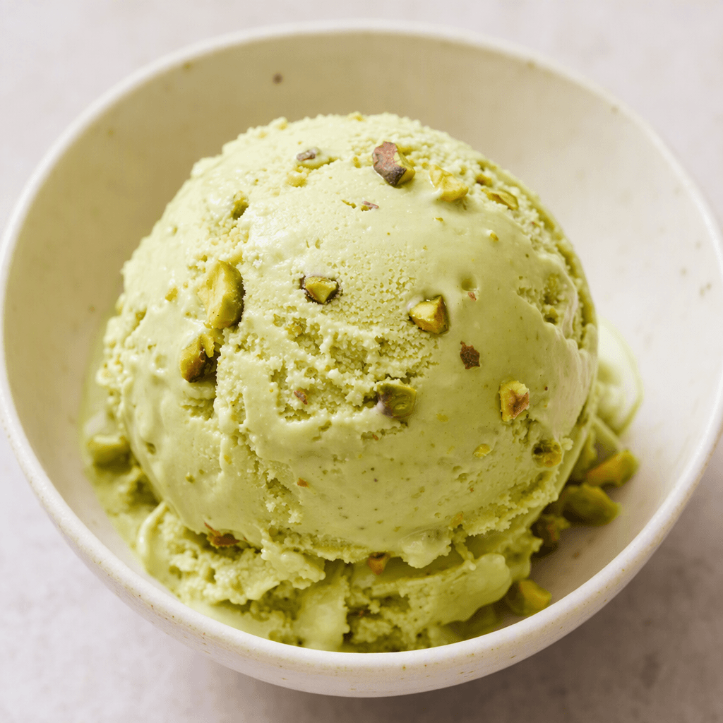 A smooth, creamy pale-green scoop of pistachio ice cream, dotted with small pieces of pistachio and served in a simple bowl.
