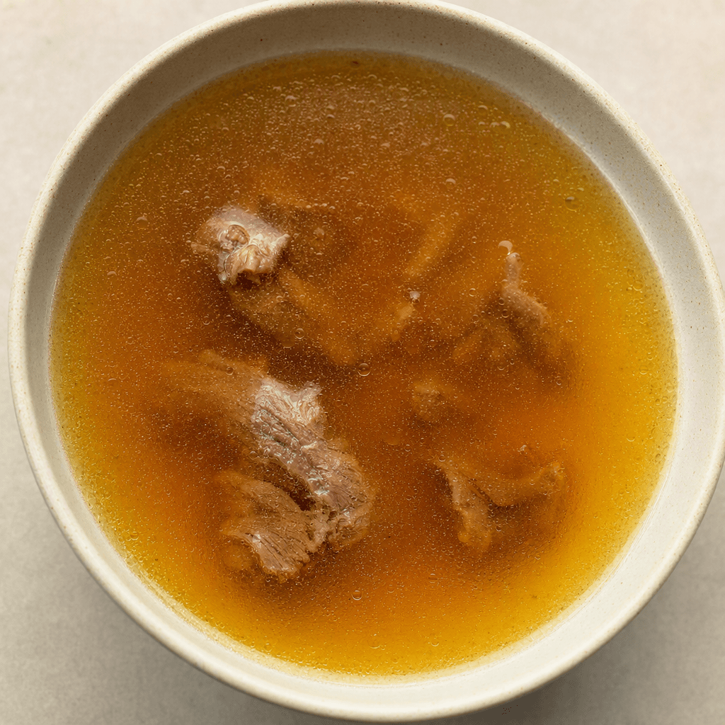 A bowl of translucent golden-brown broth with a light sheen on the surface and a few small pieces of meat visible.