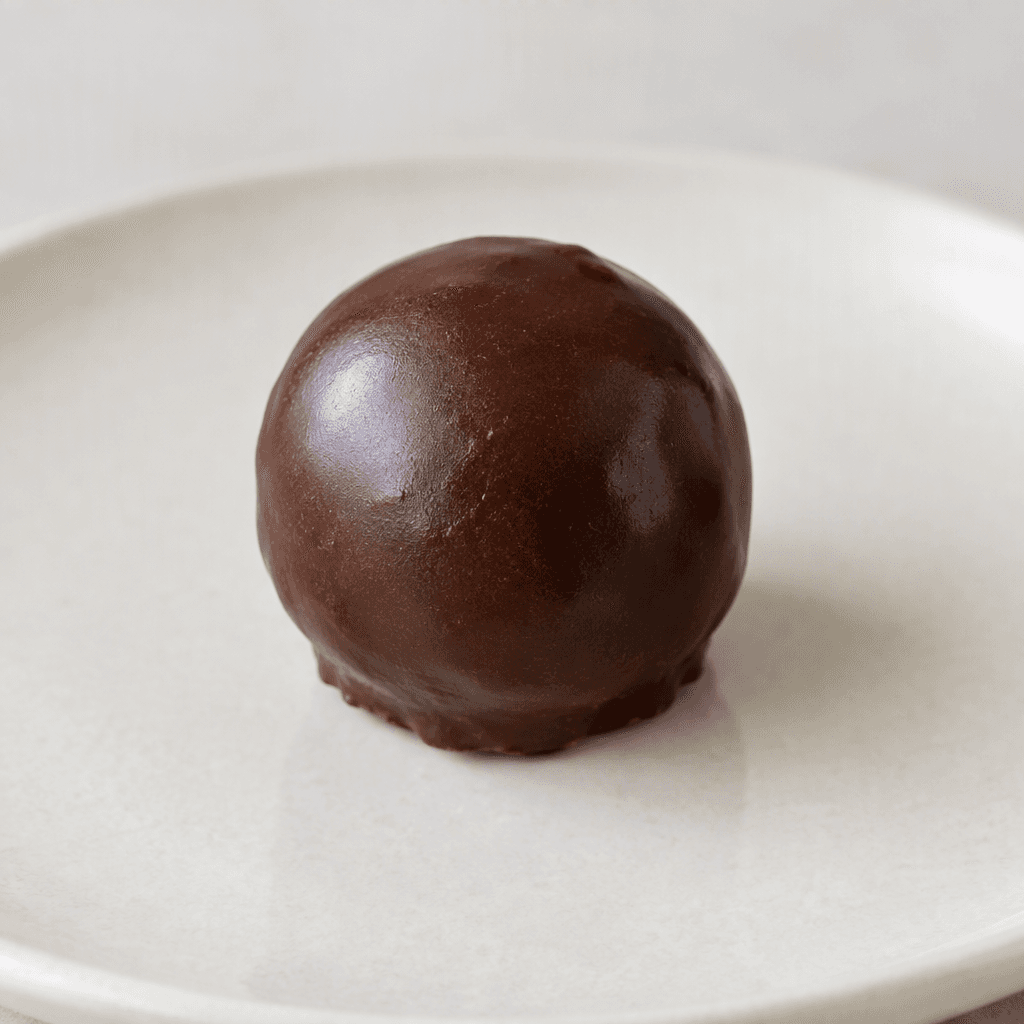 A small, round chocolate ball with a smooth, dark-brown coating, shown on a plain plate.