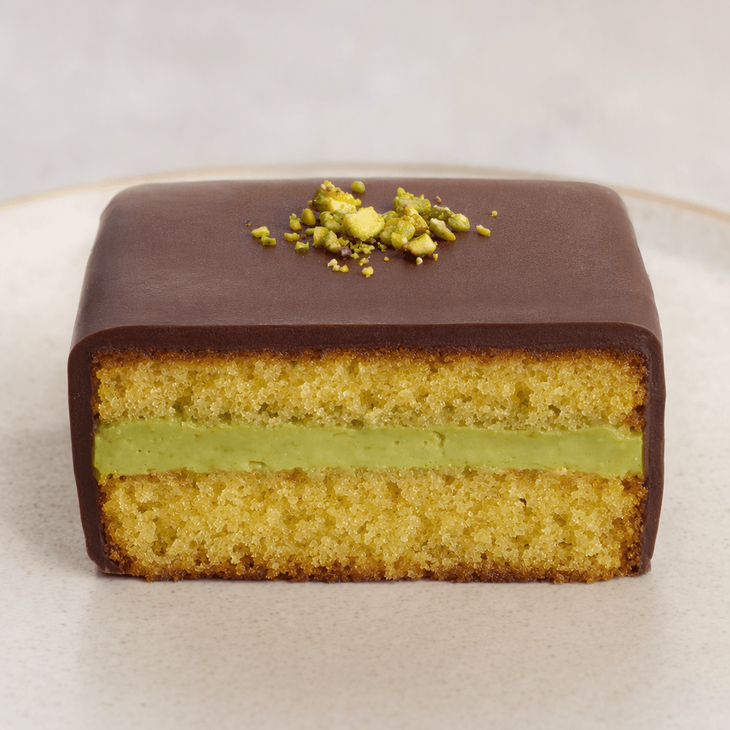 A small rectangular cake bar with a soft golden-brown sponge, a visible pale-green pistachio cream layer, and a smooth dark chocolate coating, with a few green pistachio crumbs on top.