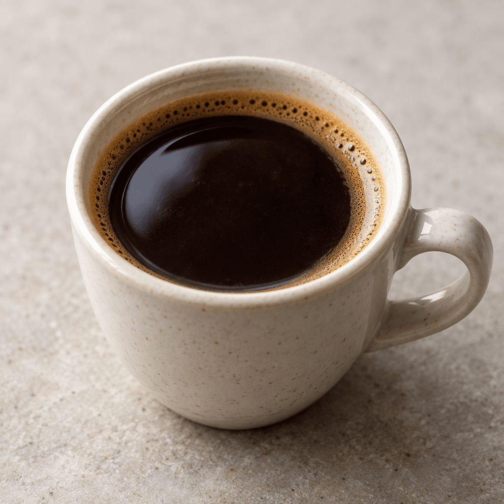 A small cup of deep brown coffee with a glossy surface and a faint, thin tan foam around the edge.