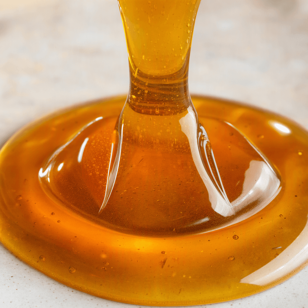A smooth, translucent golden-amber syrup forms a glossy, slowly flowing stream and a small pool.