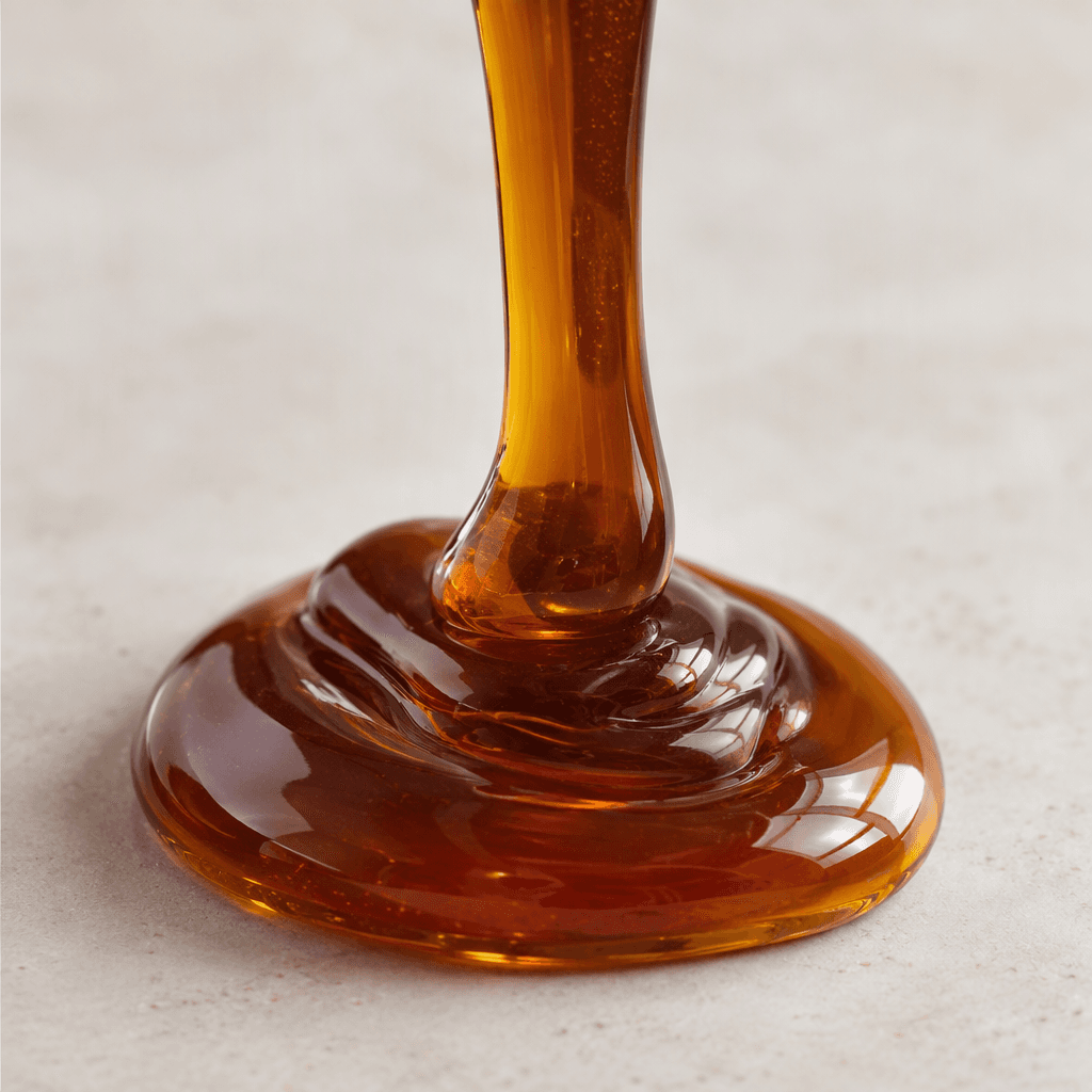 A thick, glossy, translucent dark-amber syrup forms a slow ribbon and a small pool on a plain surface.