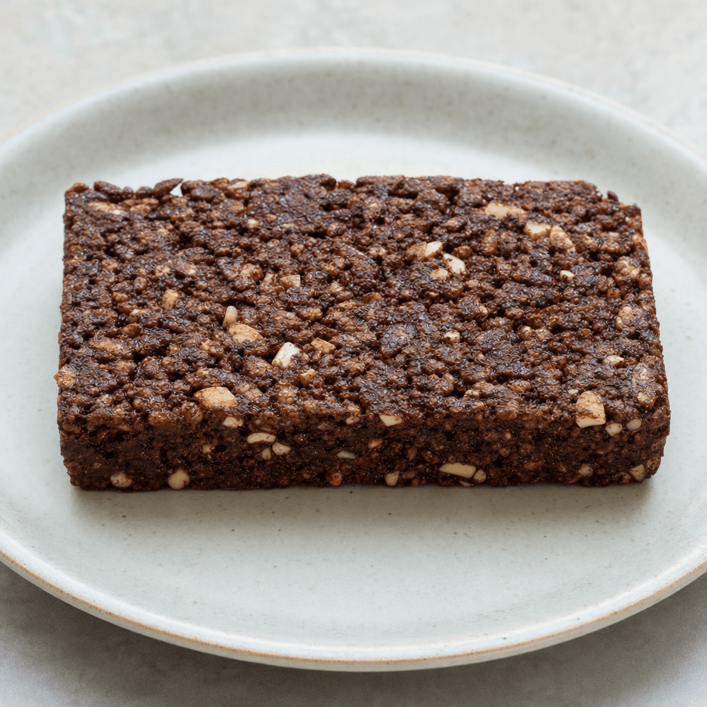 A rectangular dark chocolate cereal bar with a rough, compact surface, visible cereal grains and small pale inclusions, shown whole on a plain plate.