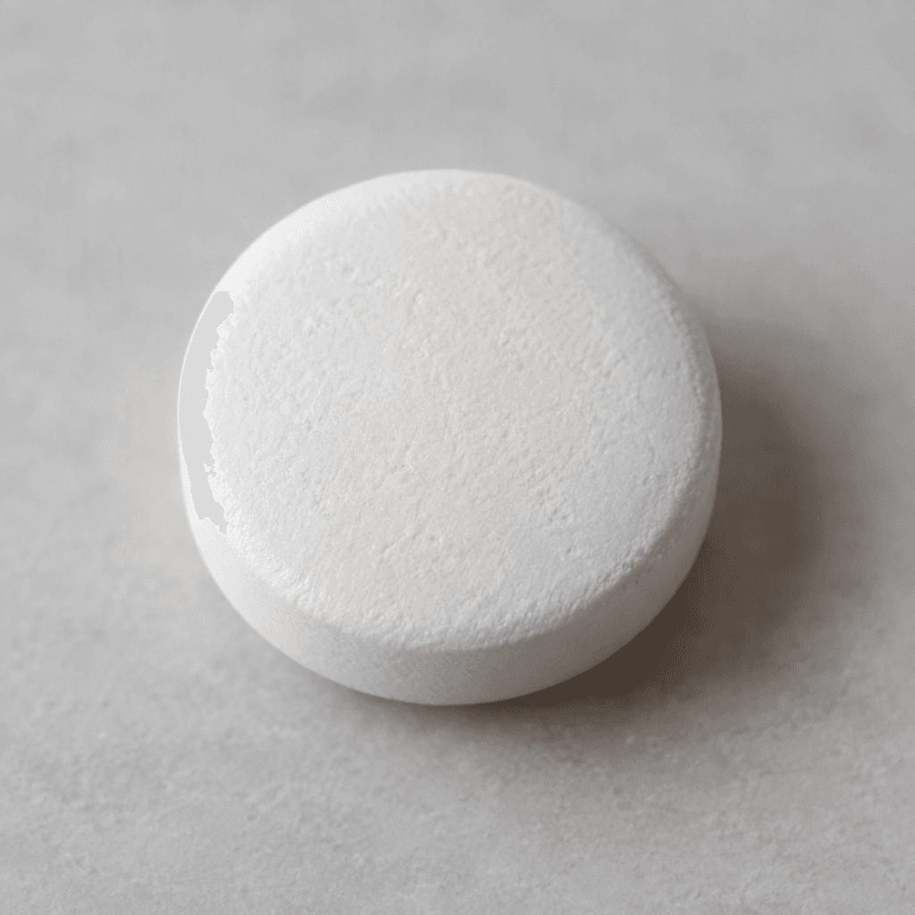 A small, smooth, white, round tablet with a flat face and slightly rounded edges rests on a plain surface.