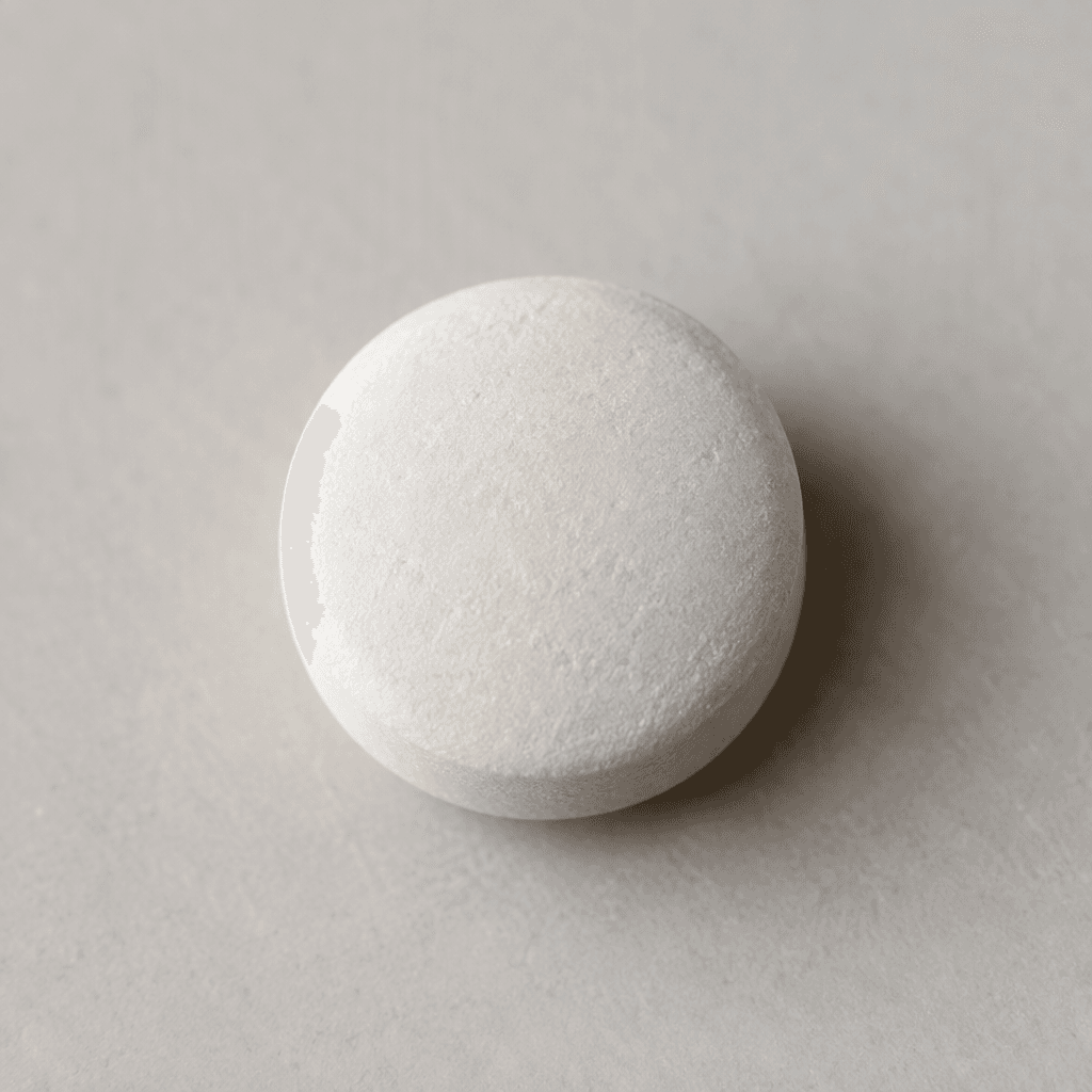 A small, round, white tablet with a smooth, slightly matte surface and gently rounded edges rests on a plain surface.