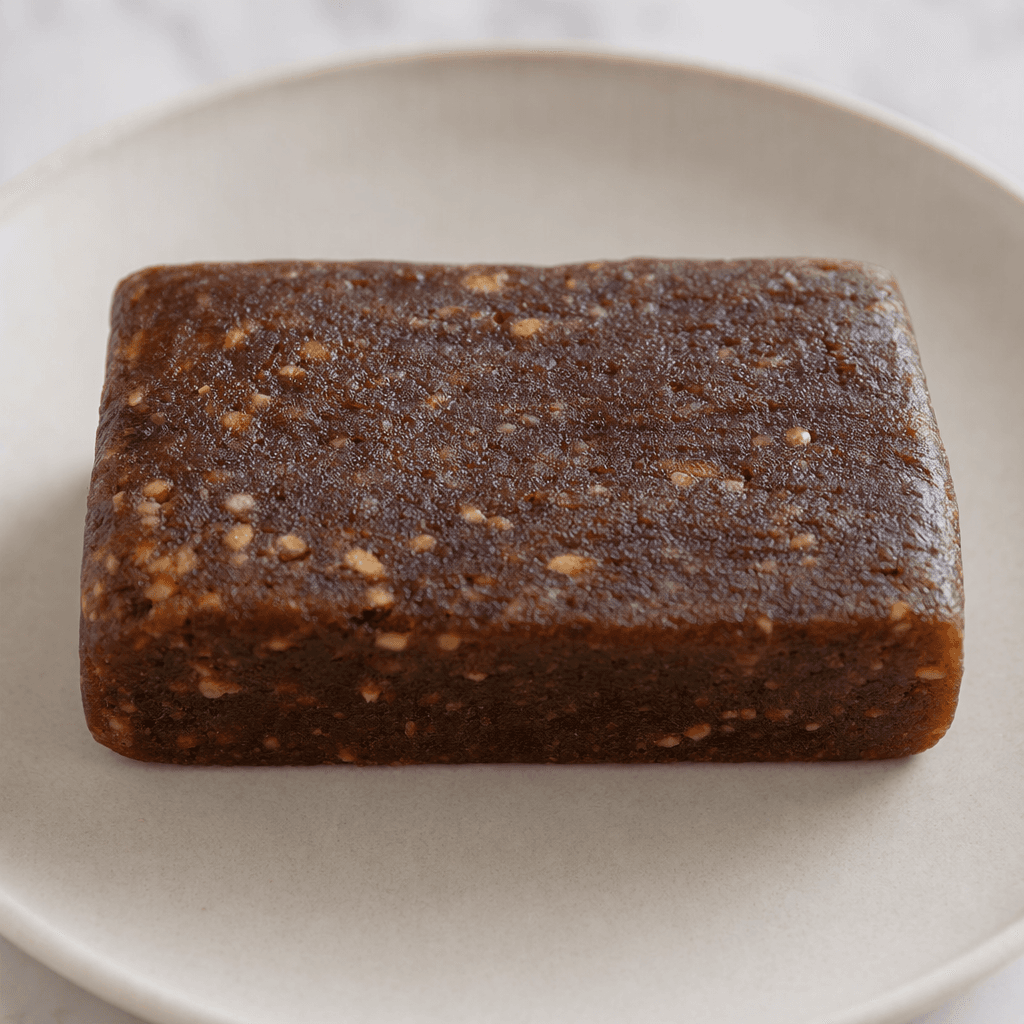 A compact rectangular dark-brown bar with a dense, slightly uneven surface and a few small lighter-brown flecks, shown on a plain plate.
