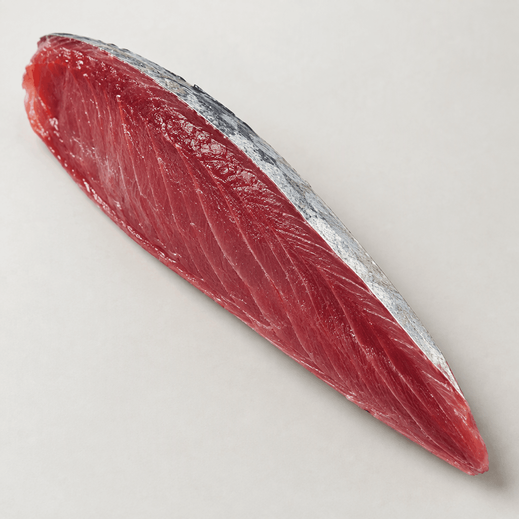 An elongated, tapered bonito fillet with moist, firm deep reddish-pink flesh and fine muscle lines, shown with a narrow strip of silvery-gray skin along one edge.