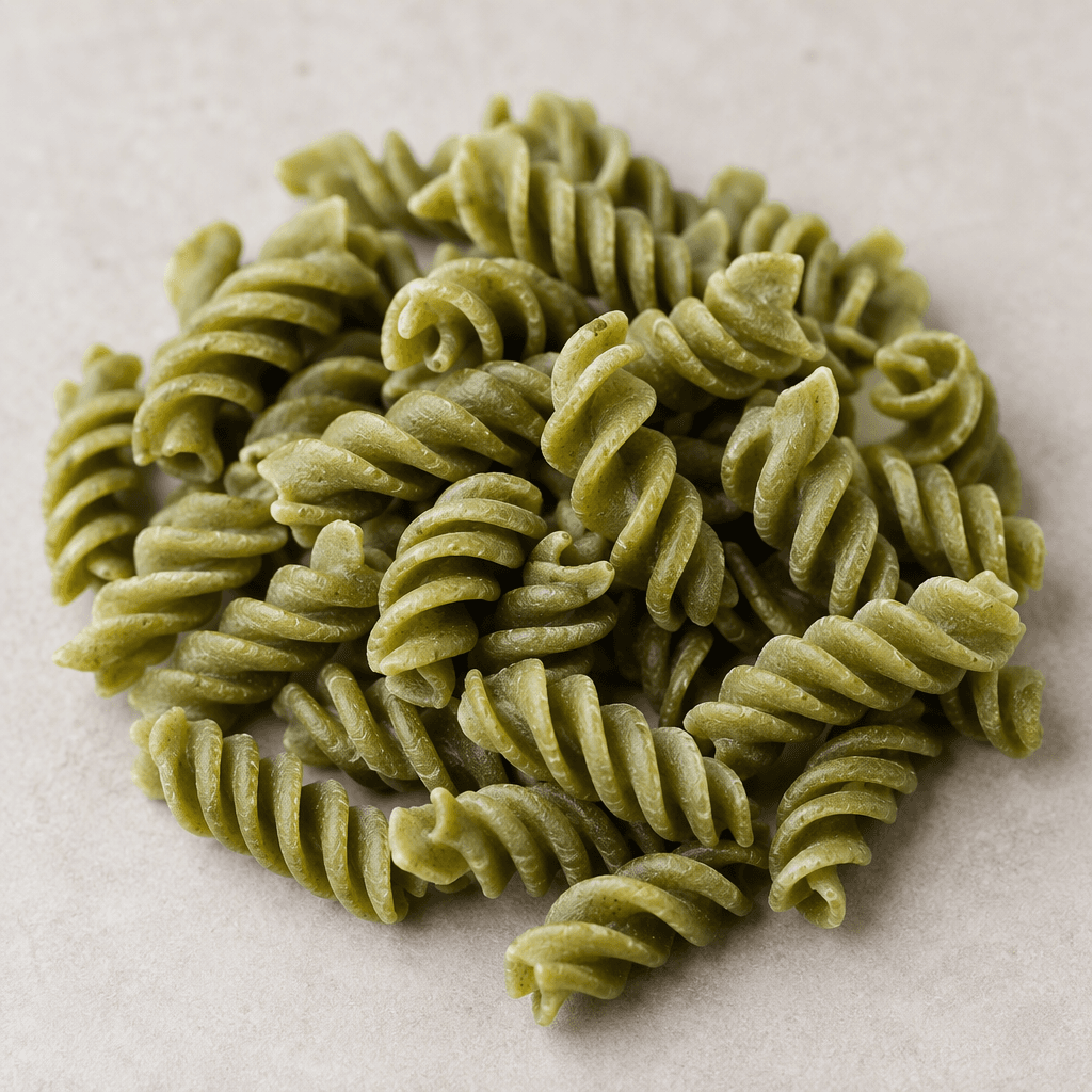 Short, dry spiral pasta pieces with ridged twists and a muted pea-green colour, scattered loosely on a plain surface.