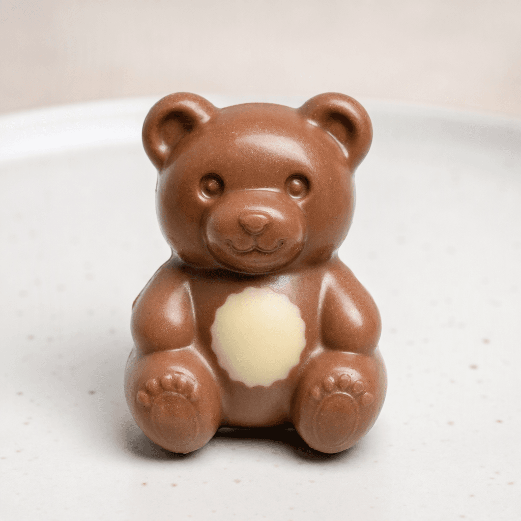 A small, glossy, light-brown bear-shaped chocolate with a pale cream-colored patch on its belly, shown on a plain plate.