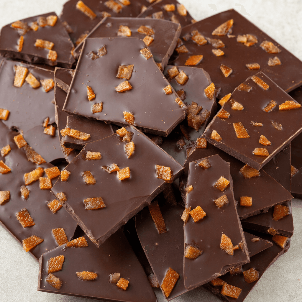 A dark-brown chocolate bar is broken into uneven squares, with small amber-orange pieces of candied peel visible across its smooth, slightly glossy surface.