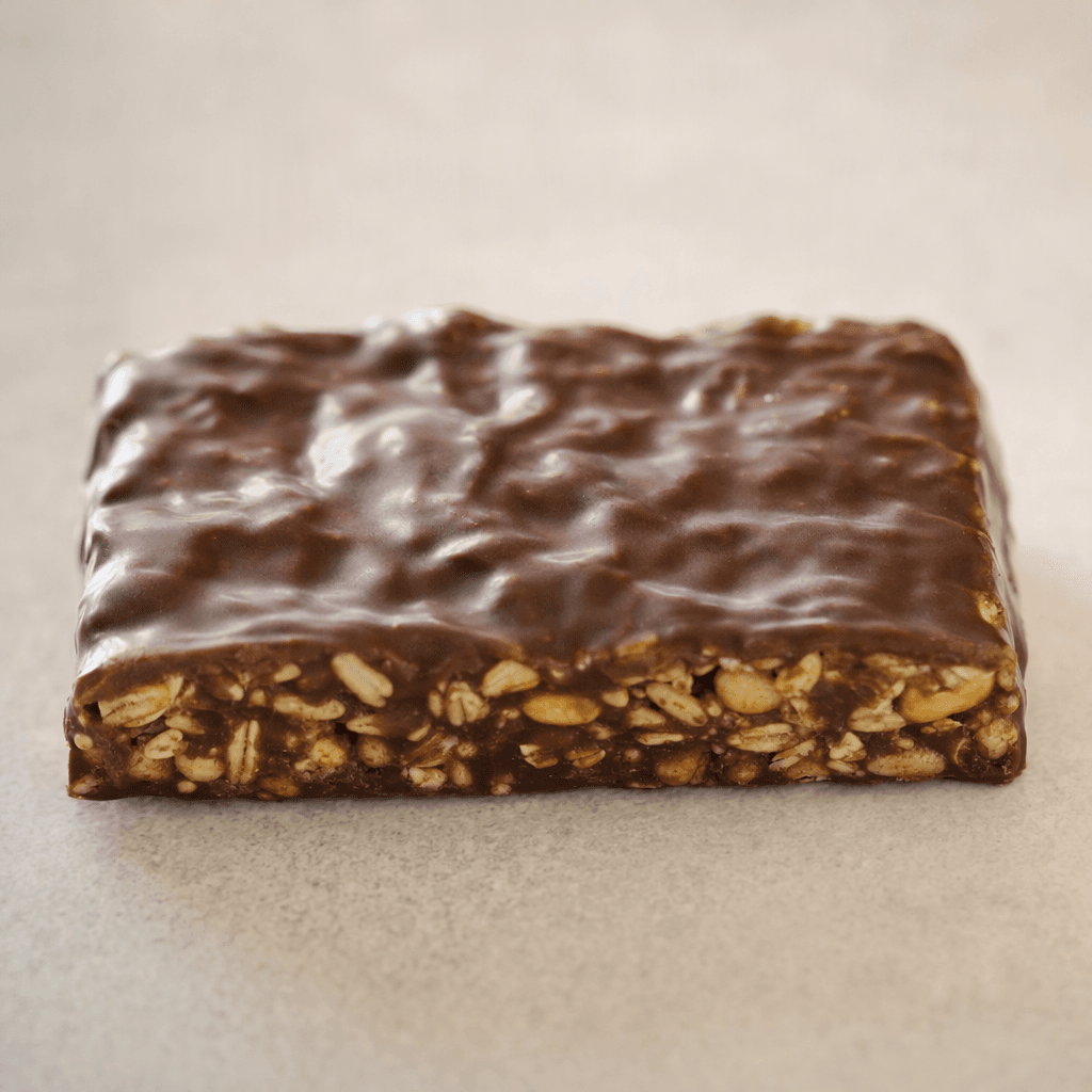 A compact rectangular cereal bar with a dark brown chocolate coating and a slightly uneven surface, with small cereal pieces visible along the edges.
