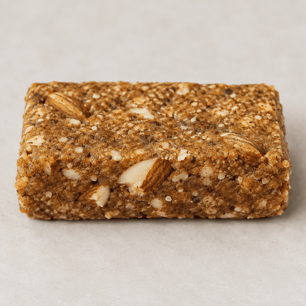 A compact rectangular protein bar with a matte golden-brown surface, slightly uneven edges, and visible almond pieces and fine grainy flecks throughout its dense, firm-looking texture.