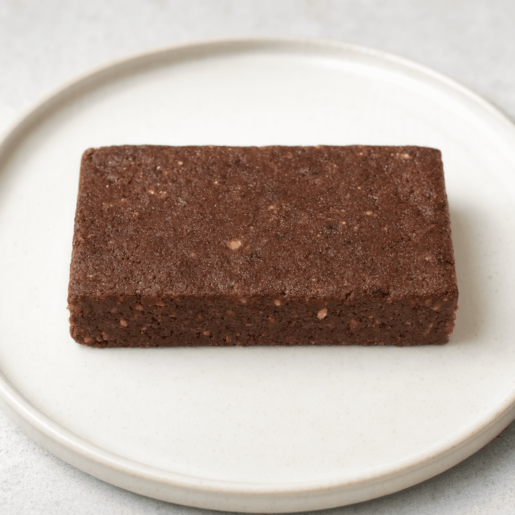 A compact rectangular chocolate-brown protein bar with a slightly uneven, matte surface and dense-looking texture, resting by itself on a plain plate.