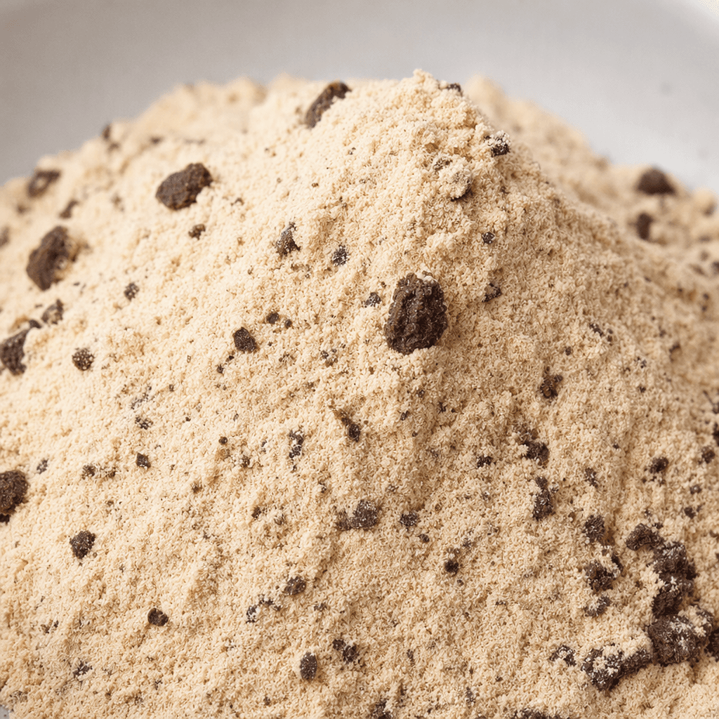 A mound of fine, light beige protein powder with scattered darker brown cookie-like flecks and a soft, slightly grainy texture.